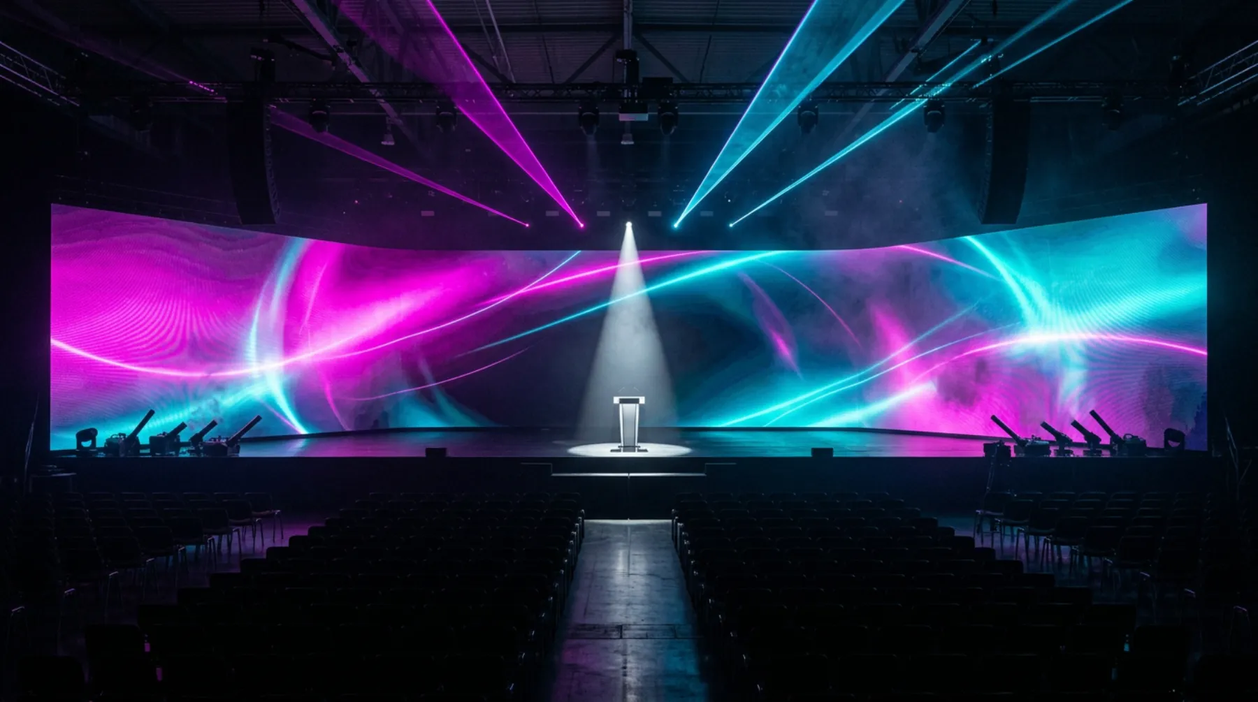 A huge empty keynote stage: a lone podium in a white spotlight before a giant curved screen of magenta and cyan light, lasers overhead, confetti cannons at the edges and empty seats in the dark.